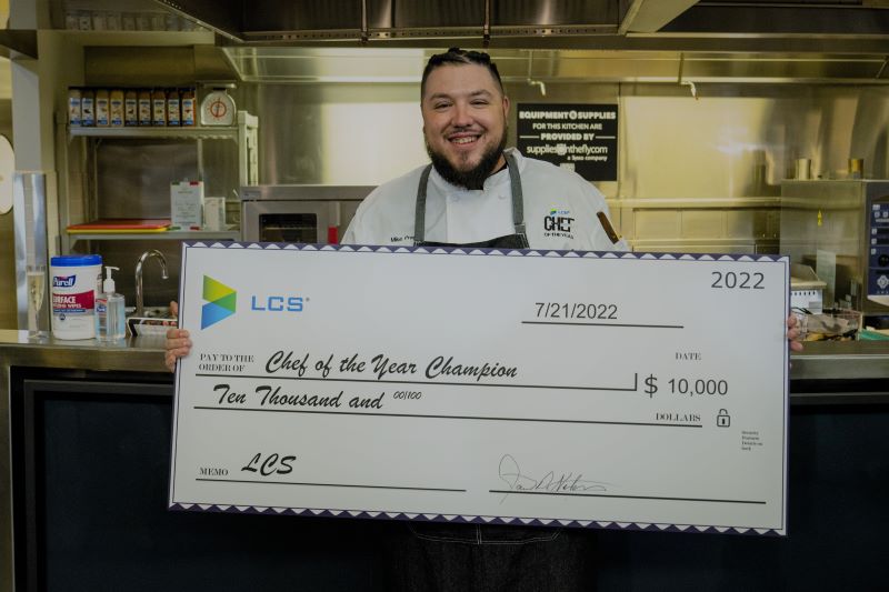 A professional kitchen, a chef is smiling at the camera holding a cheque that reads Chef of the Year Champion, $10,000