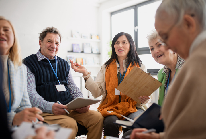 Middle aged woman with a clipboard leads a meeting with four others.