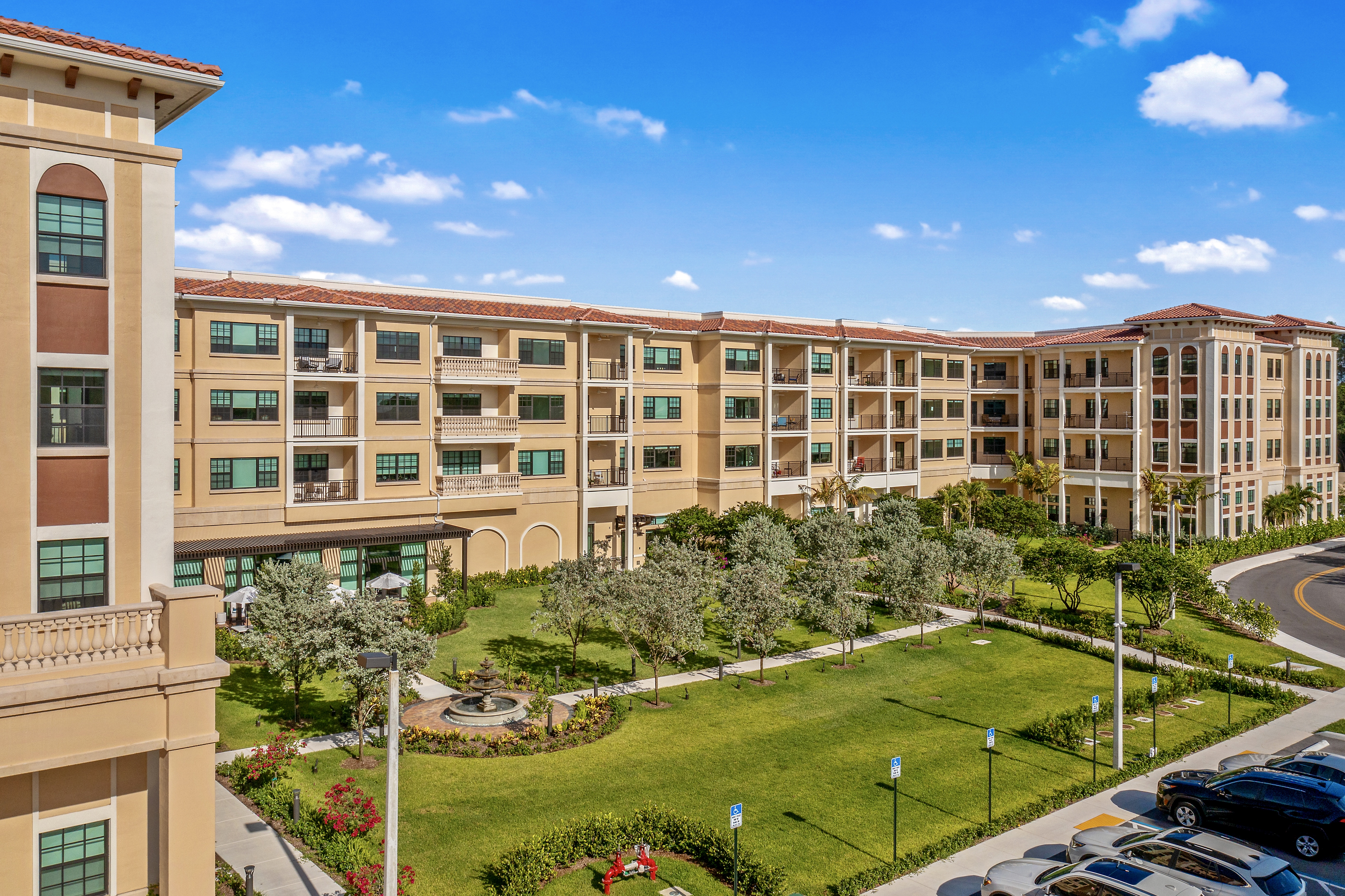 A large four-story building with a green space, trees, and a fountain, along with a car park with cars parked.