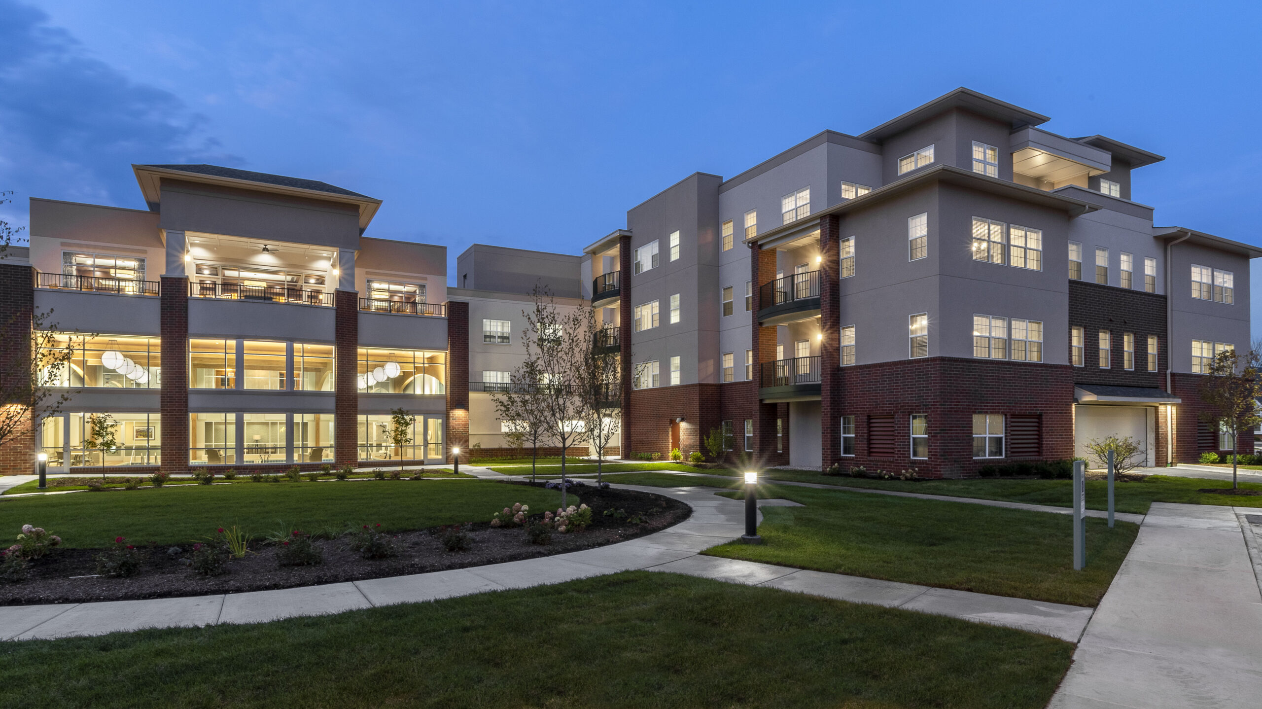 An outside setting at dusk of a large lit-up four-story building, Wyndemere, on grounds with paths and green spaces.