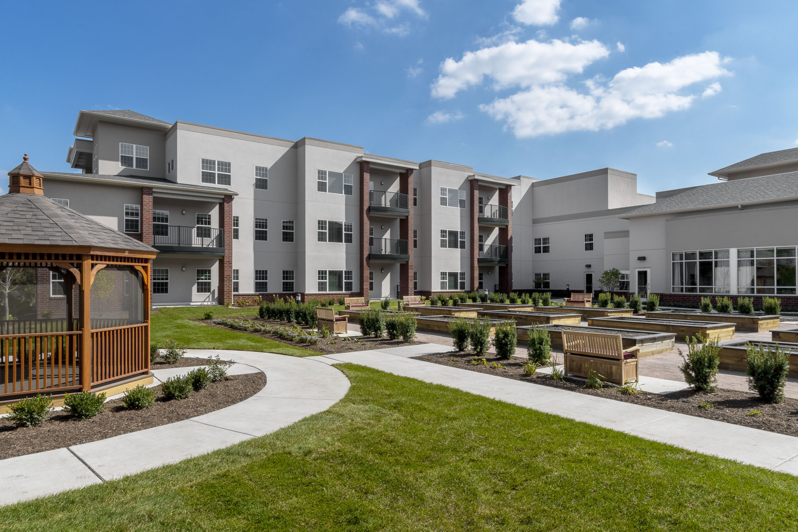 An outside view of a large three-story building, Wyndemere, behind an expanse of raised flower beds, pathways, benches, and a garden shelter.