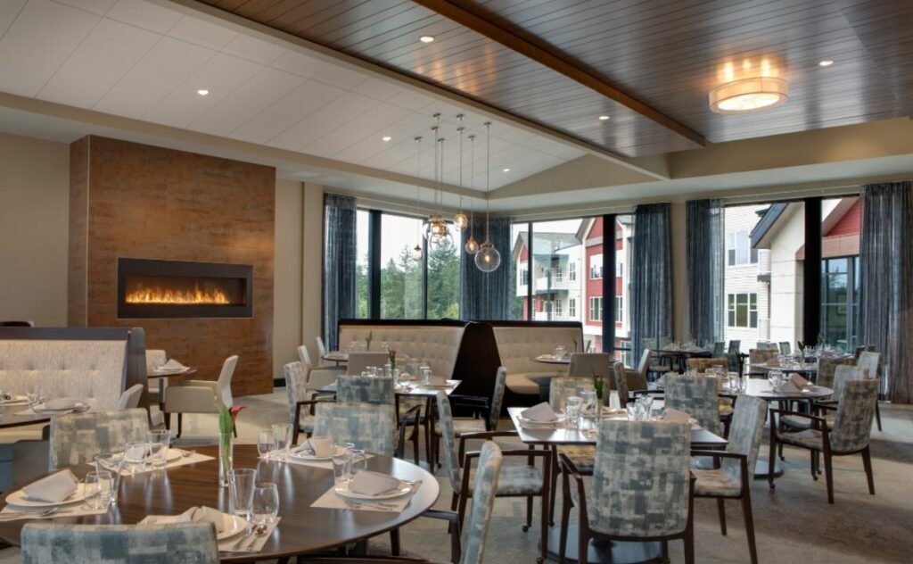 A large dining room with circular and square tables set for dinner with tableware. A wall-mounted electric fire is on in the background.