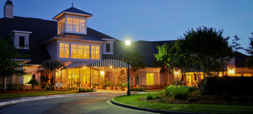 An outside view of a building at dusk with warm, inviting lights, and a road outside surrounding a green island with trees and bushes.