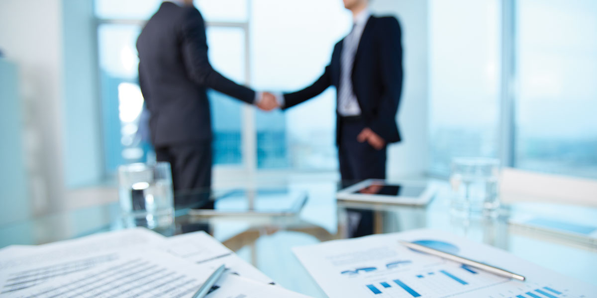 An office location, in the background out of focus two men in business suits shaking hands, in the foreground in softer focus is a desk with papers of charts and columns and two pens.