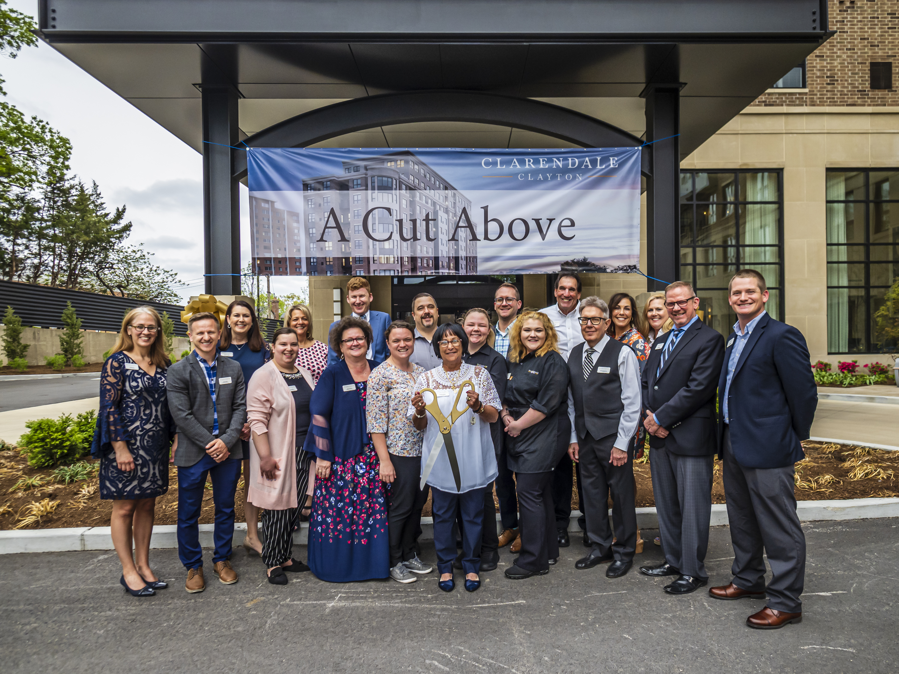 A group of people standing outside the Clarendale Clayton building smiling at the camera, one woman holding ceremonial scissors in front of a banner saying A Cut Above.