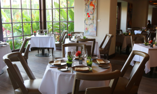 A communal dining room with circular and square tables with seating, laid for dinner, in front of a bay window.