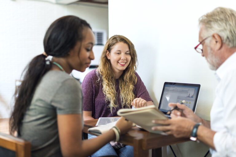 An older man and two younger women sitting at a table with books and laptops. The man is pointing at something in a book and the two women are smiling at it.